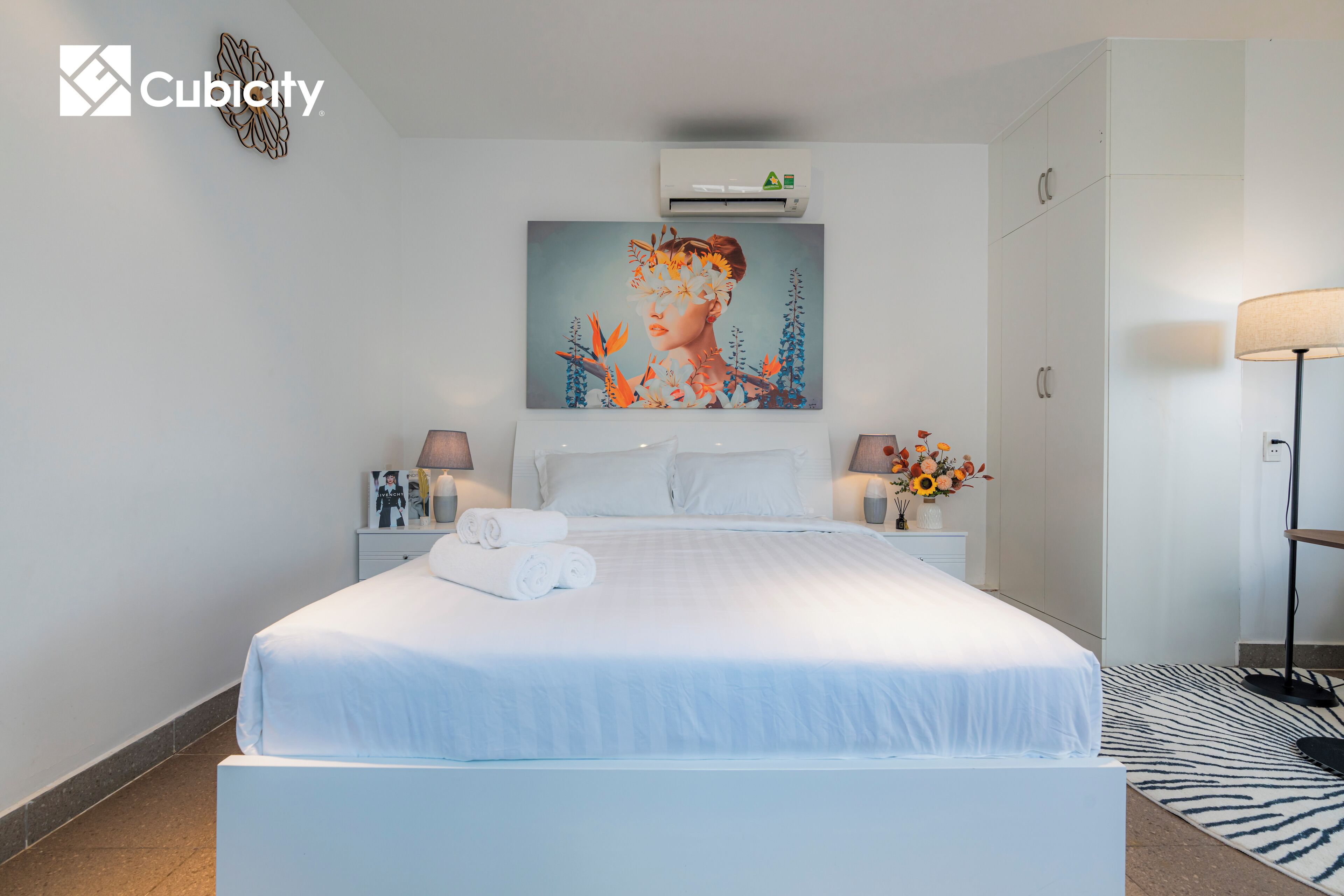 A hotel room with a large bed, a painting on the wall, an air conditioning unit, a lamp, and a zebra-patterned rug.