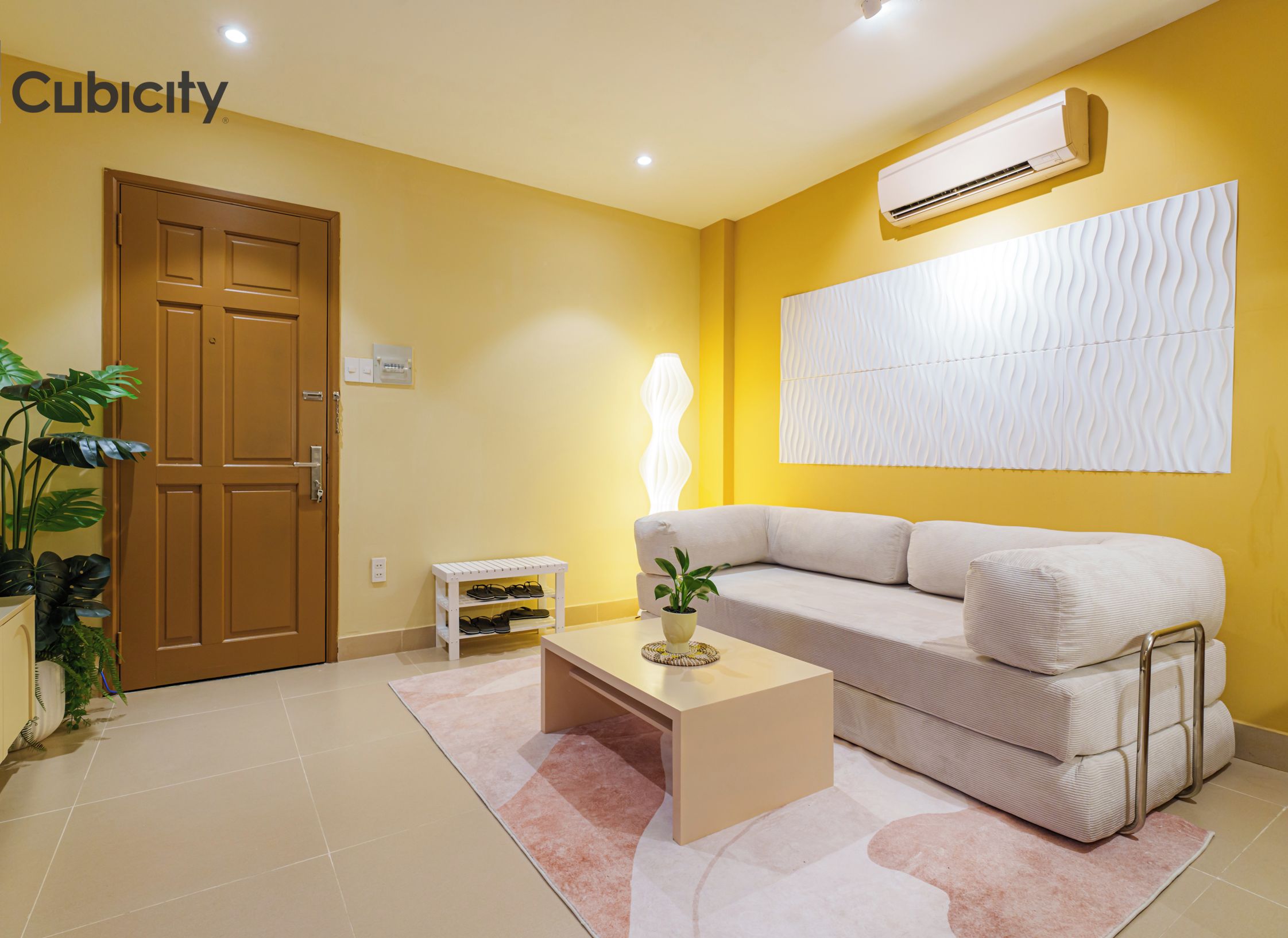 A living room with a beige sofa, a coffee table, a potted plant, a shoe rack, and a door.