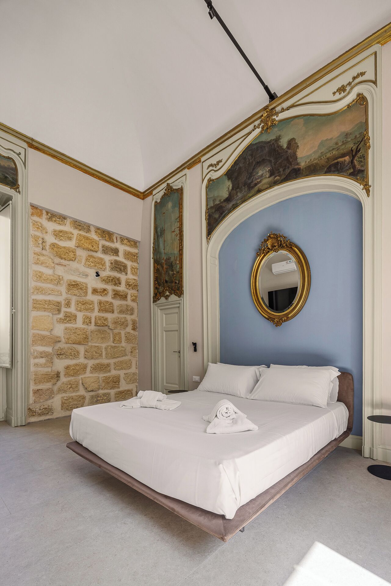 A bedroom with a stone wall, a bed with white linens, a large mirror, and a door.
