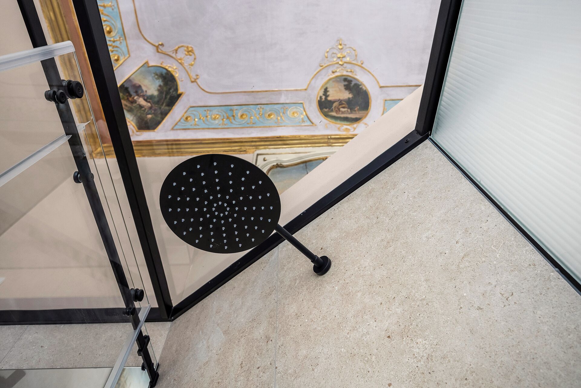 Canto de um quarto com divisória de vidro, um objeto circular preto com furos e piso de azulejos.