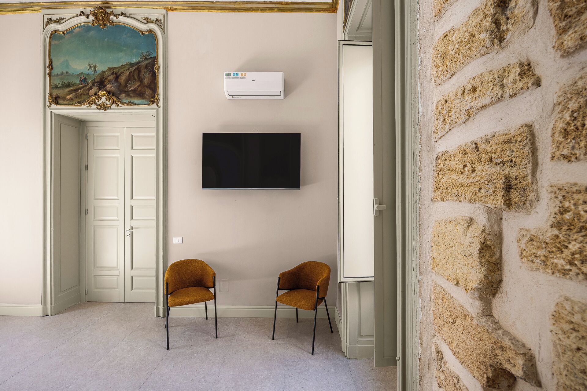 A room with a white door, a mounted TV, two orange chairs, and a stone wall.