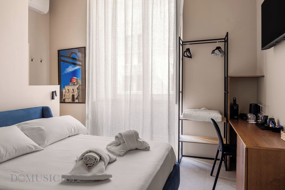 Habitación de hotel con cama, escritorio, silla, televisor y ventana con cortinas traslúcidas.
