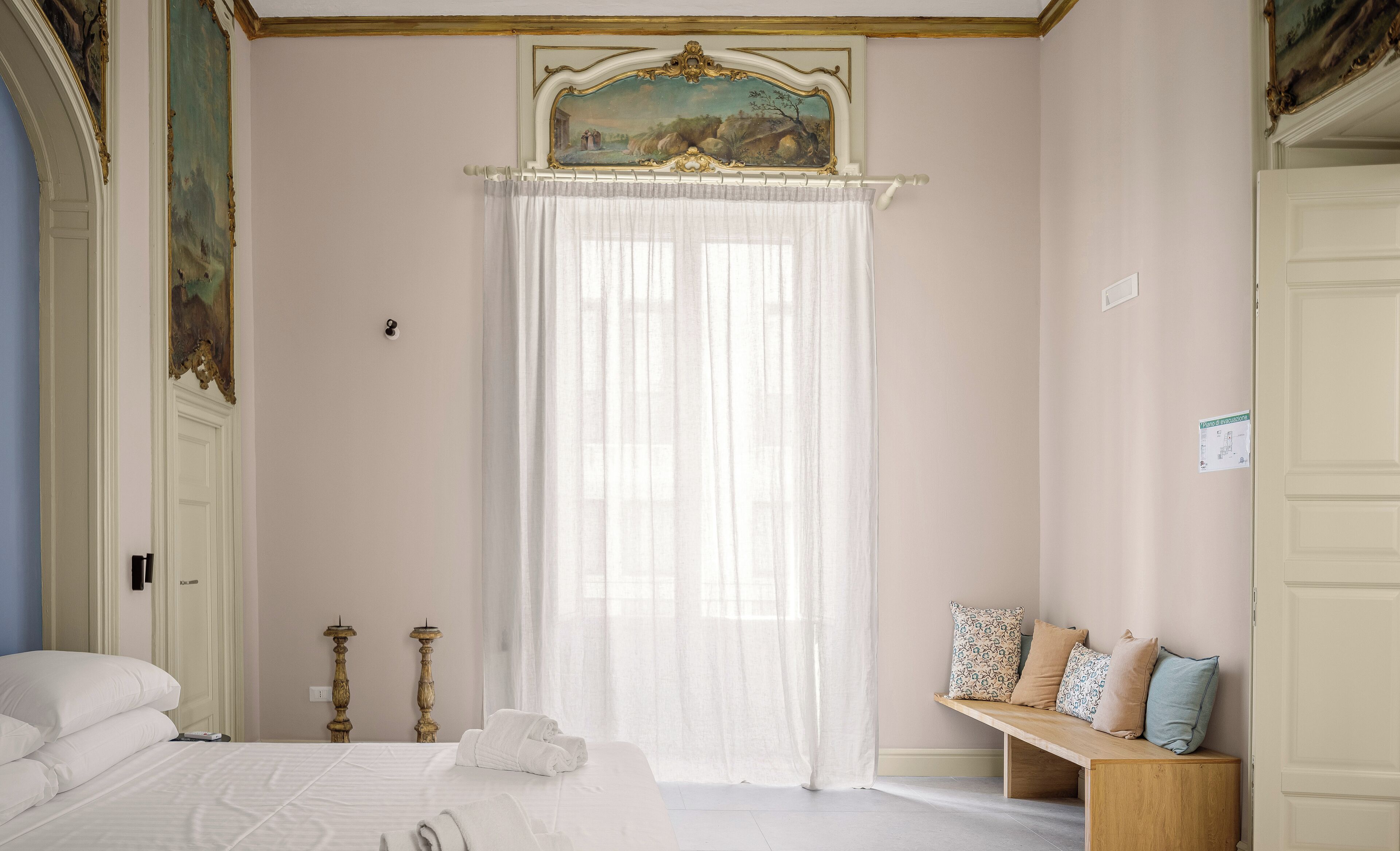 A bedroom with a bed, a bench with pillows, a painting above the window, and a door with a sign.