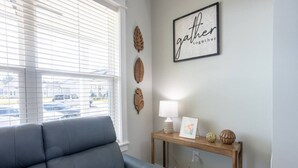 A living room with a grey sofa, a wooden console table with a lamp, framed artwork, and decorative leaves on the wall. - Lofty Lakeside Cottage – 2 BR, 2.5BA, Sleeps 8! (Greenwood)