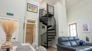 A modern living room with a gray leather sofa, a dining table with a vase and placemats, a spiral staircase, and artwork on the walls. - Lofty Lakeside Cottage – 2 BR, 2.5BA, Sleeps 8! (Greenwood)