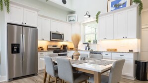 A modern kitchen with white cabinets, stainless steel appliances, a dining table with chairs, and a window with a view of the outdoors. - Lofty Lakeside Cottage – 2 BR, 2.5BA, Sleeps 8! (Greenwood)