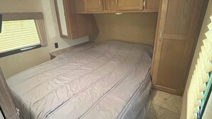 A compact RV bedroom with a bed, wooden cabinets, and a window with blinds.