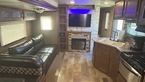 A compact RV interior with a living area featuring a leather sofa, a fireplace, and a flat-screen TV.