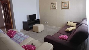 A living room with a beige sofa, a dark brown sofa, and a small ottoman. There is a television on a black stand, and two framed pictures on the wall.