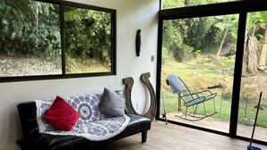 Un salon moderne avec un canapé en cuir noir, un plaid à motifs et un coussin rouge. Il est doté de grandes fenêtres offrant une vue sur la verdure, ainsi que d’un patio avec une chaise suspendue et un balai bleu.