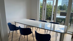 Un'area pranzo moderna con tavolo in vetro e sedie blu scuro, ampie finestre che offrono vista su vegetazione e un edificio, e un pavimento in legno.