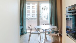 A modern kitchen with a round dining table and chairs, a hanging light fixture, and a view of a balcony with curtains.