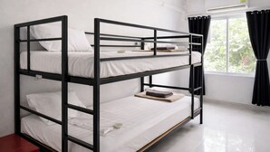 Habitación con literas, ropa de cama blanca, un pequeño armario rojo y una ventana con cortinas negras.