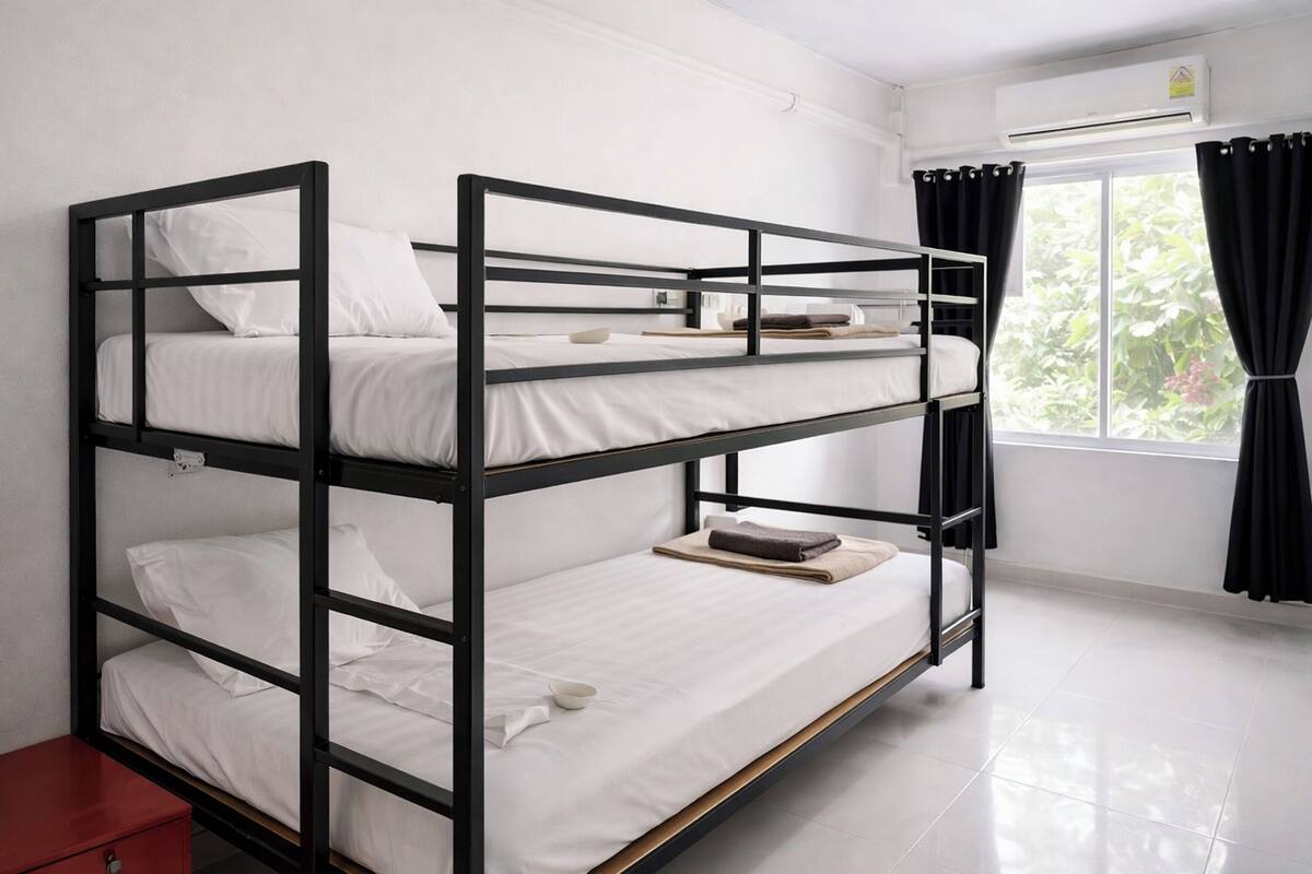 A bunk bed room with white bedding, a small red cabinet, and a window with black curtains.