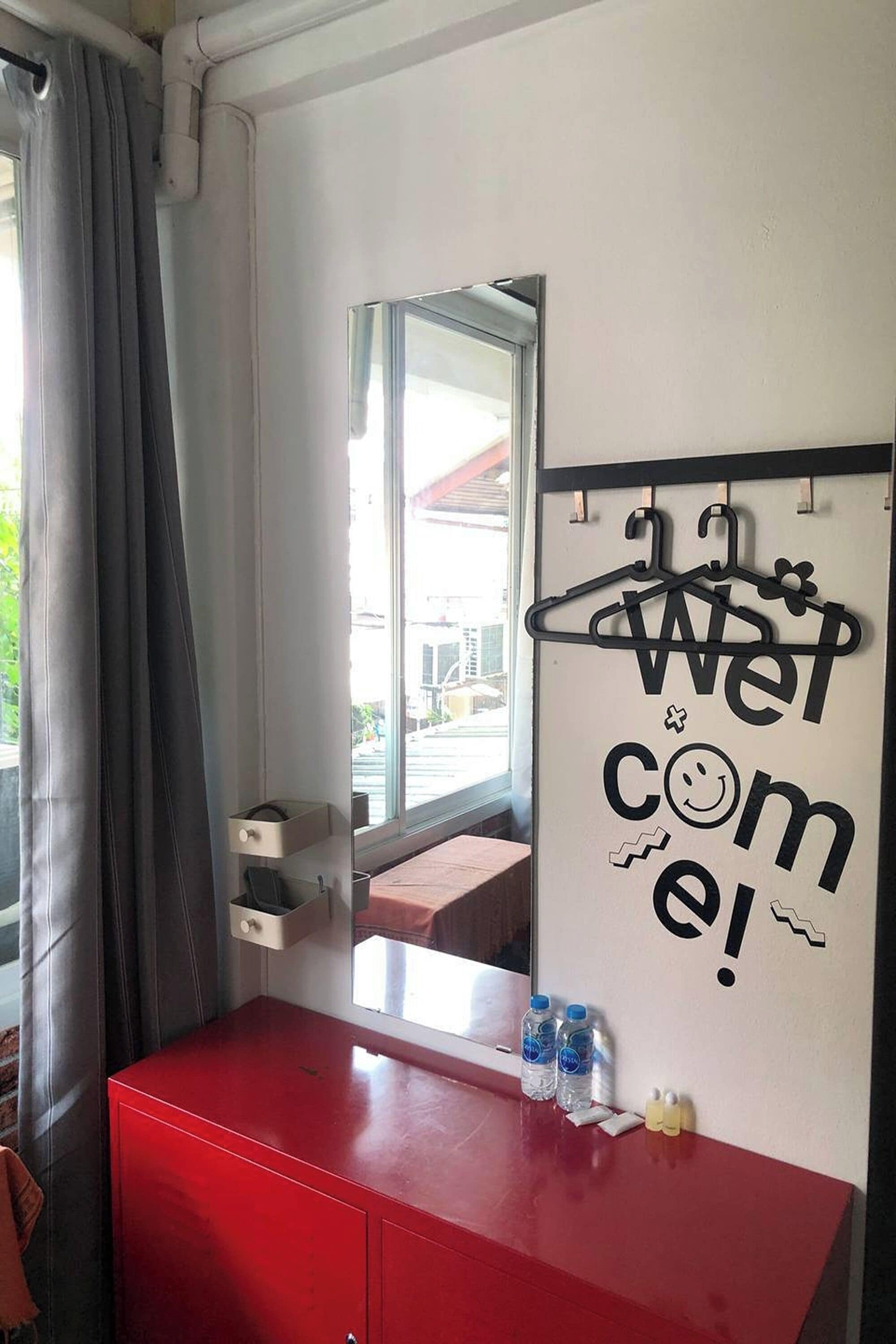 Ein Zimmer mit einem roten Schrank, einem Spiegel, einem Fenster mit Vorhängen und einer Wand mit einem 'Welcome'-Schild.