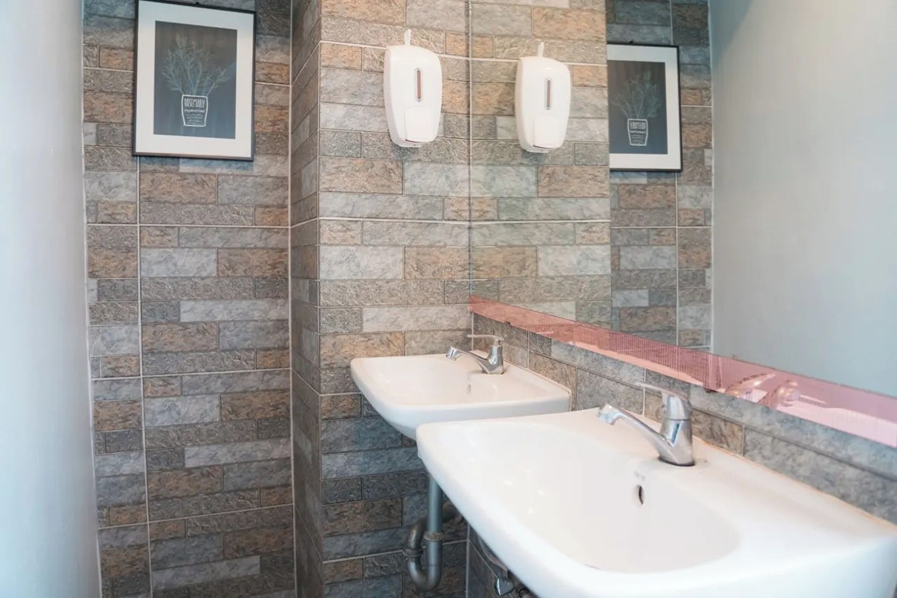 A bathroom with two sinks, a mirror, and soap dispensers on a stone-tiled wall.