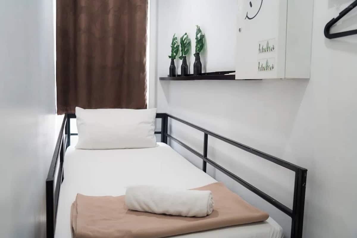 A single bed with a white mattress, a beige blanket, and a white pillow. There is a brown curtain partially drawn to the side. A black metal headboard is visible.