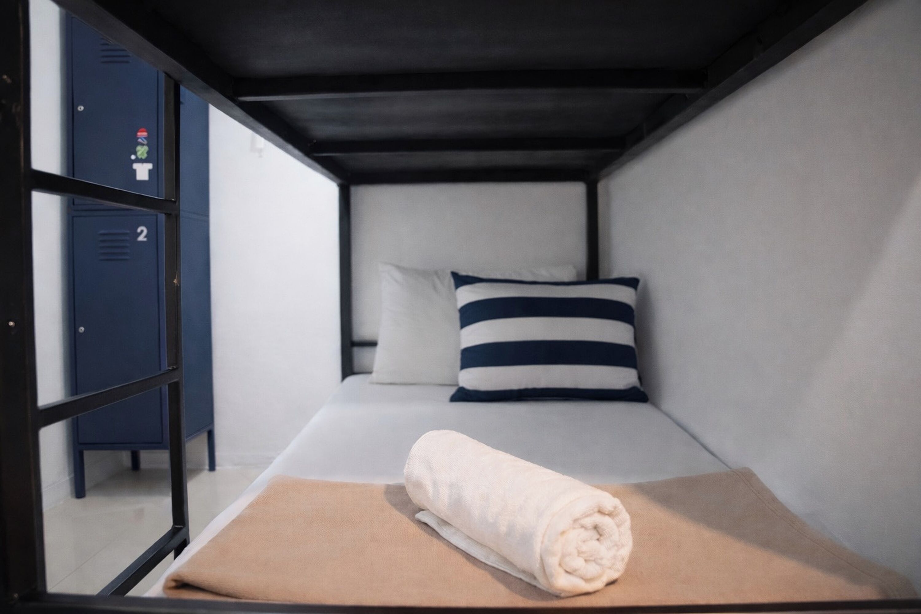Een kleine kamer met een enkel bed, een opgerolde handdoek en een gestreept kussen. In de achtergrond zijn twee kluisjes zichtbaar.