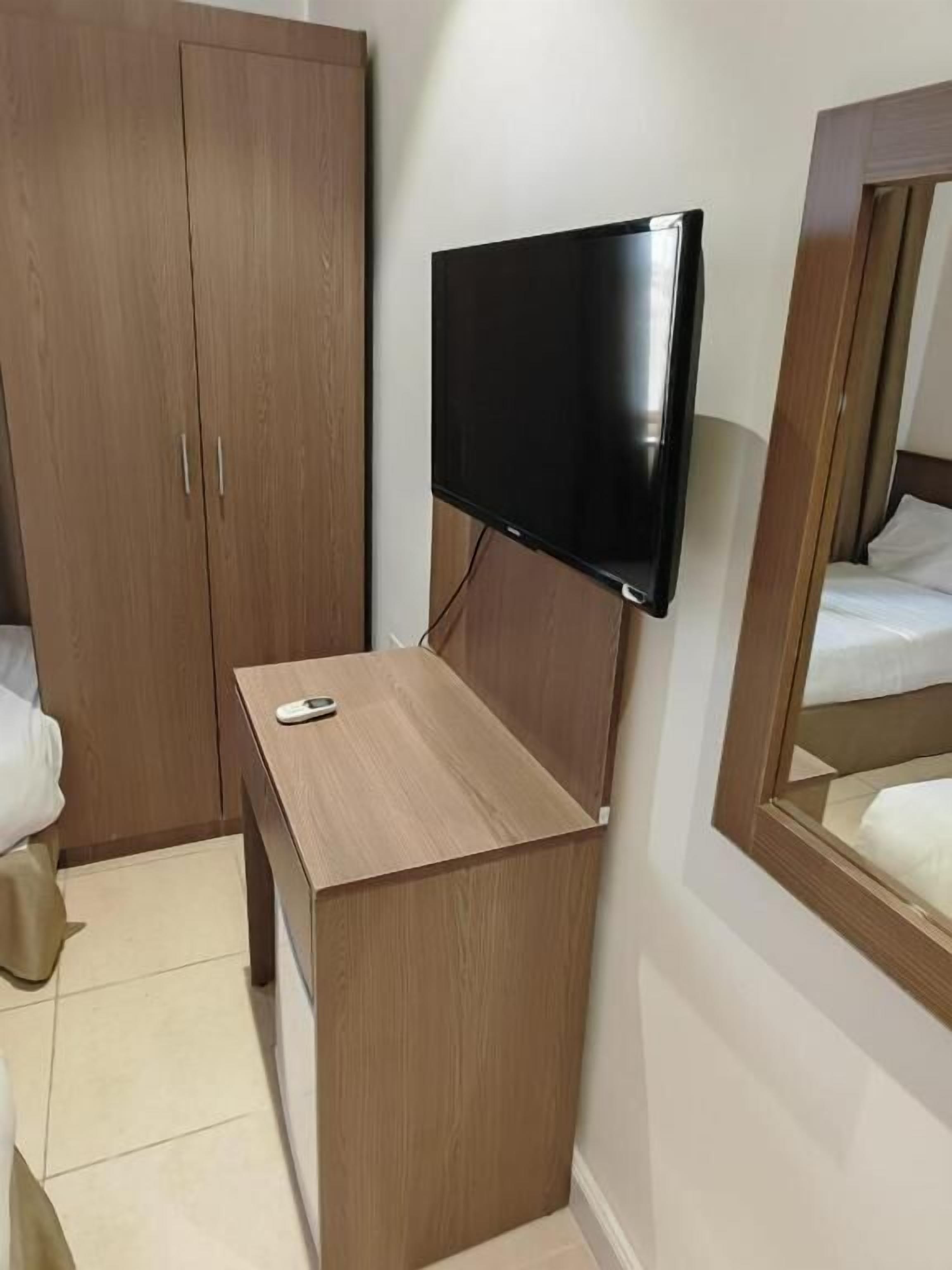 Habitación de hotel con armario de madera, mesita de noche con control remoto, televisor de pantalla plana en la pared y cama con ropa blanca.