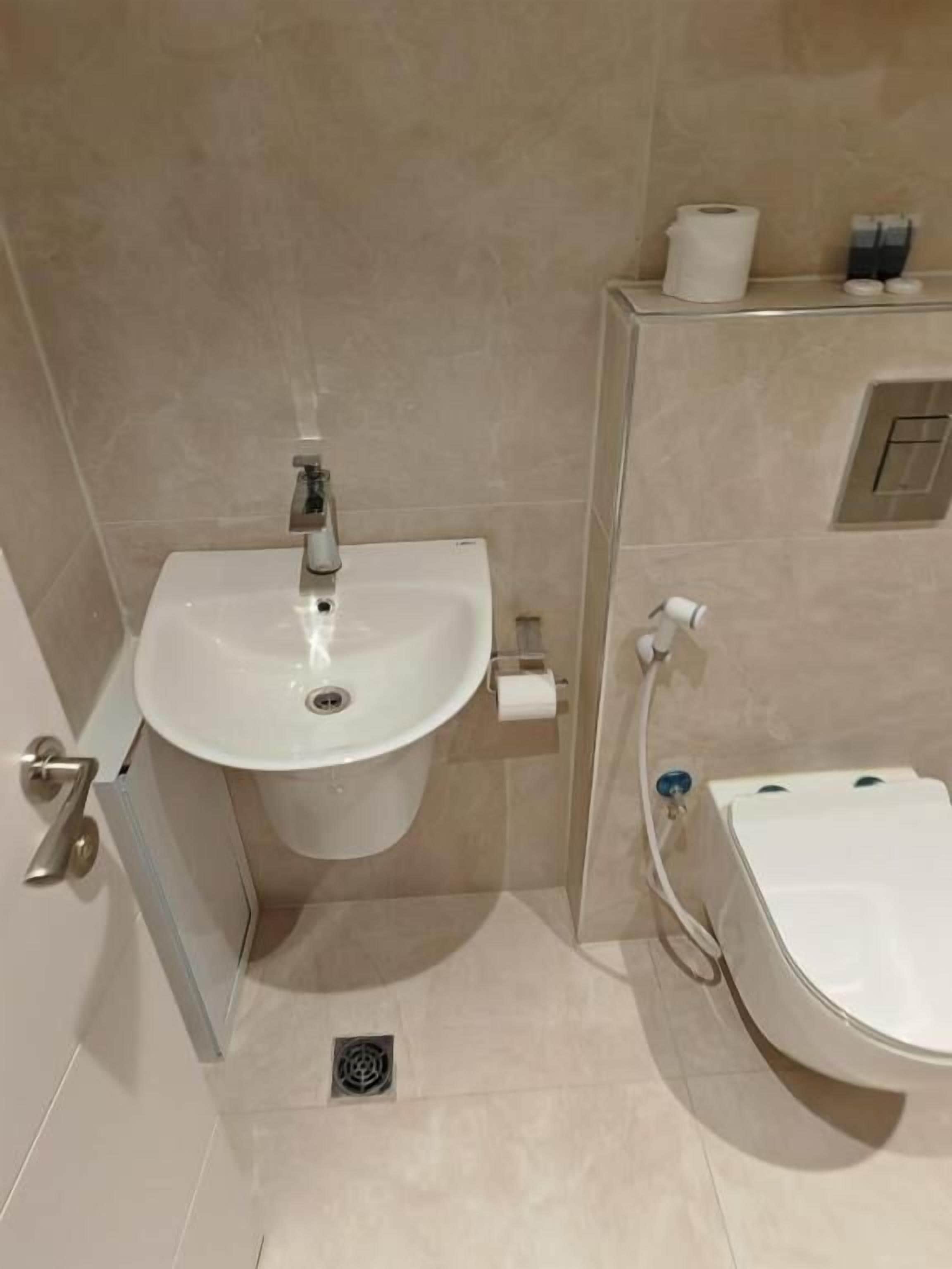Un baño con lavamanos, inodoro y bidé blancos. Hay un dispensador de papel higiénico y un pequeño cubo de basura en el suelo.