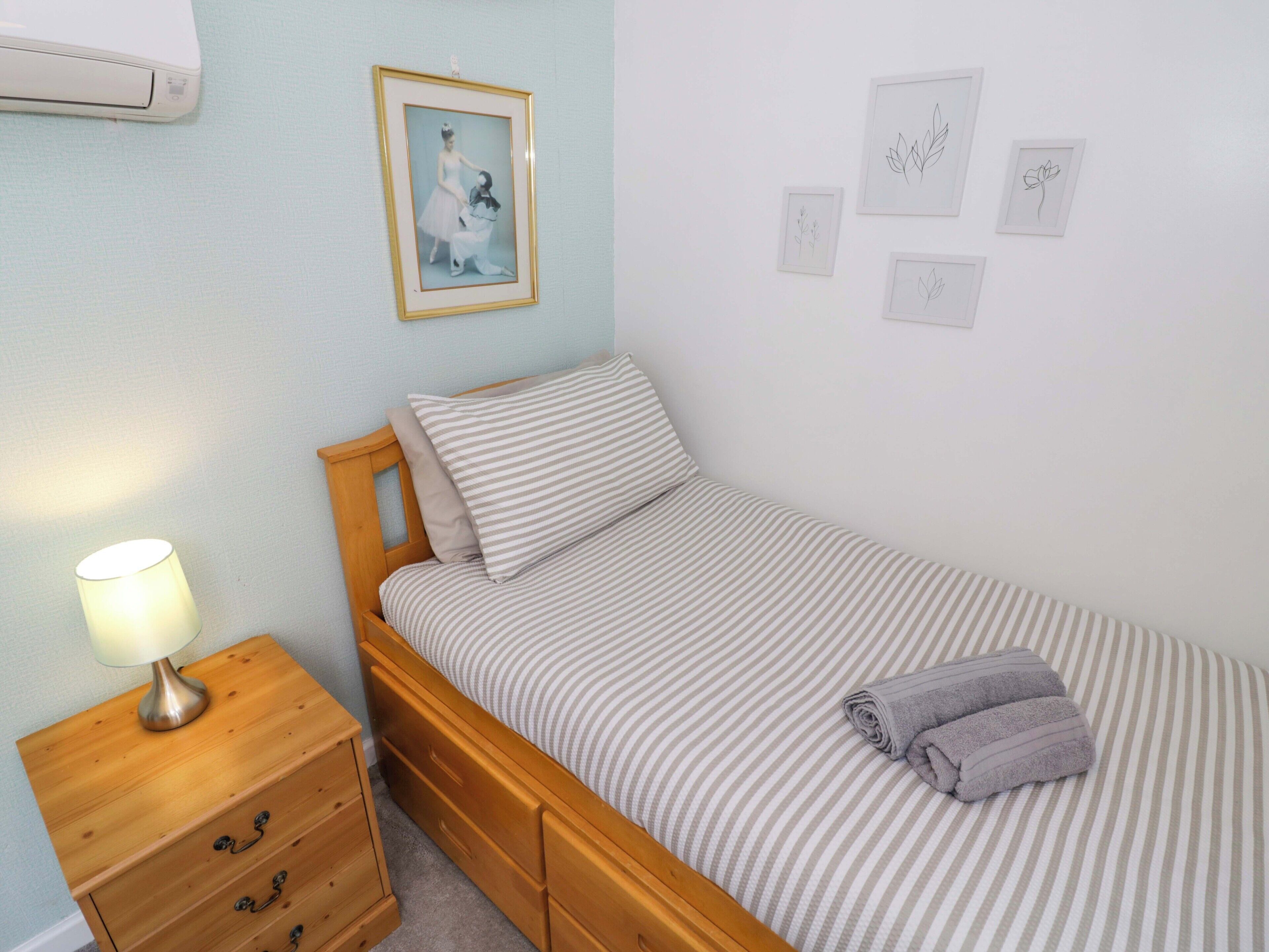 Un dormitorio con una cama individual, una mesita de noche de madera con cajones, una lámpara y un cuadro en la pared.