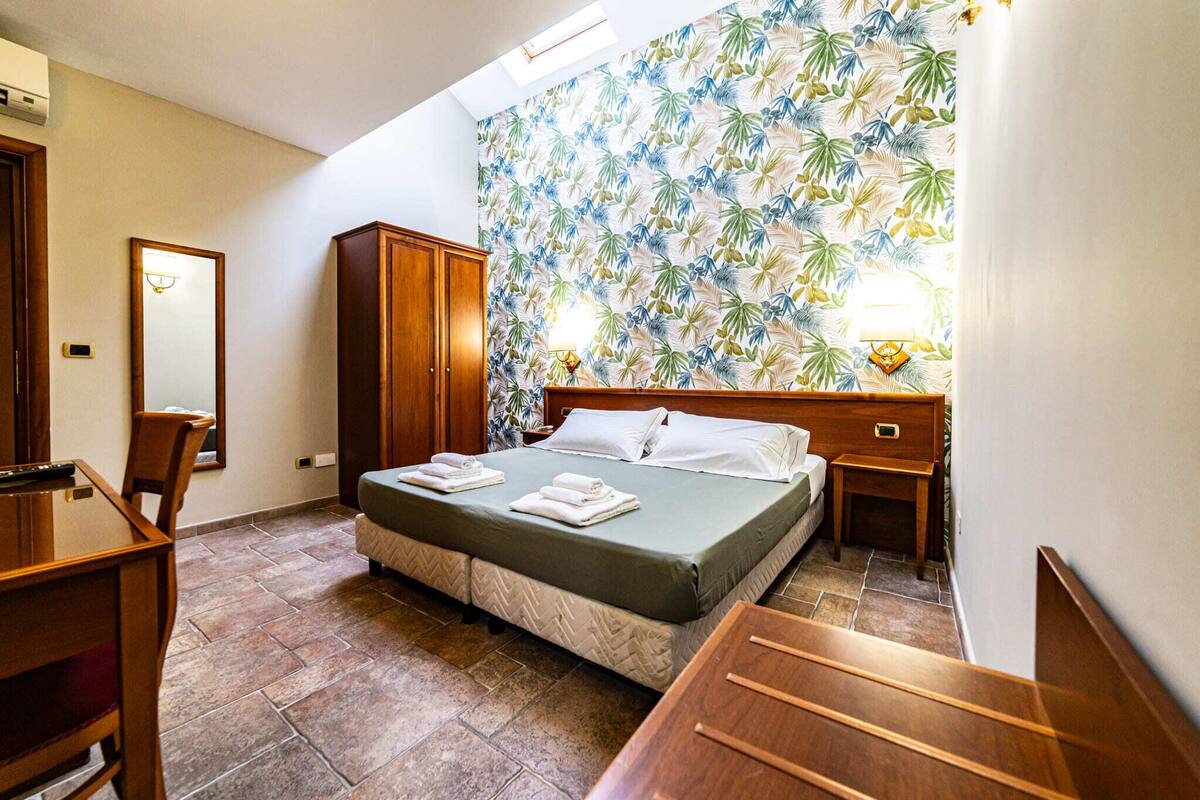 A hotel room with a bed, a desk, a wardrobe, a mirror, and a tropical wallpaper.