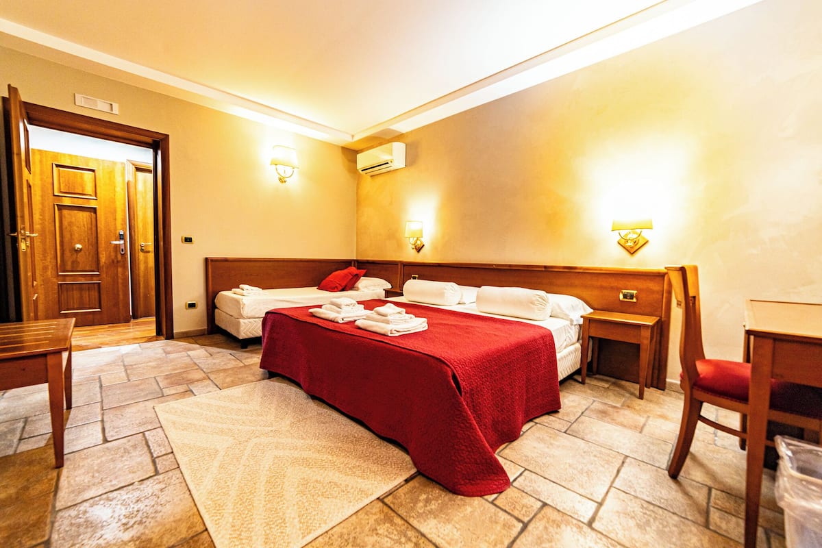 A hotel room with a bed, a desk, a chair, a wooden door, and a wall-mounted air conditioner.