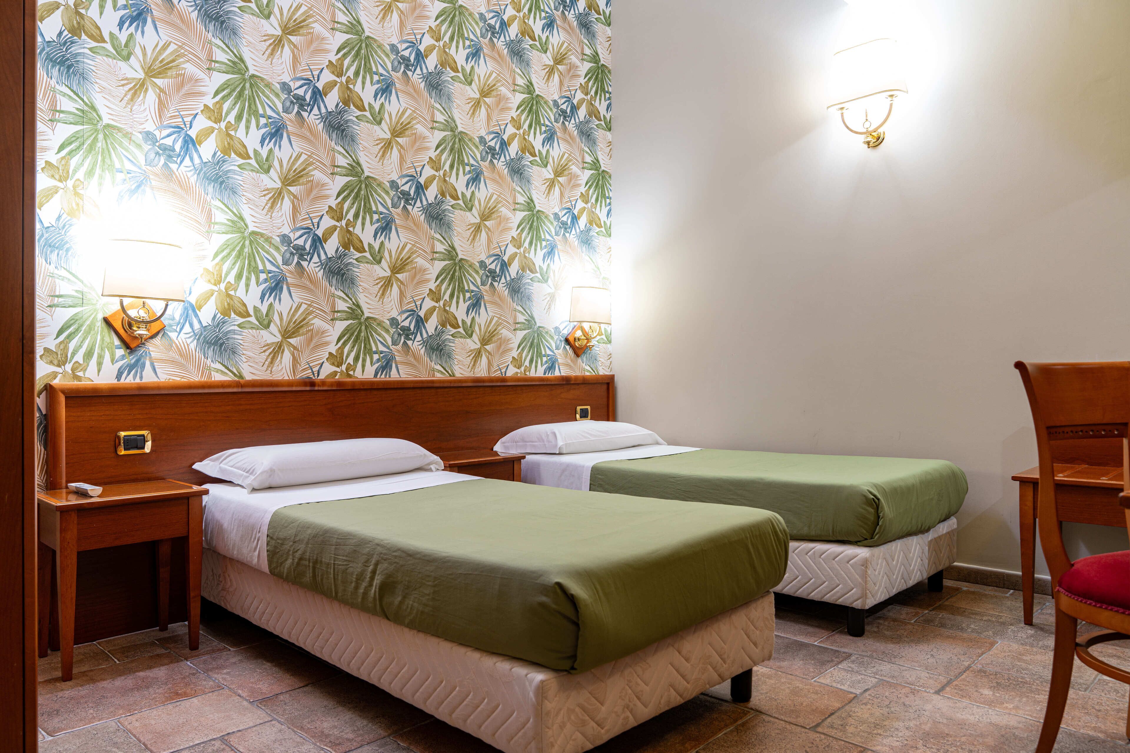 A hotel room with two single beds, a wooden headboard, a nightstand with a lamp, a chair, and a wall with a tropical leaf pattern.