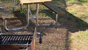 Un área de picnic con una larga mesa de madera y bancos, rodeada por una cerca de alambre y árboles.