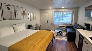 Una habitación de hotel compacta con cama, sofá, kitchenette pequeña y ventana con cortinas.