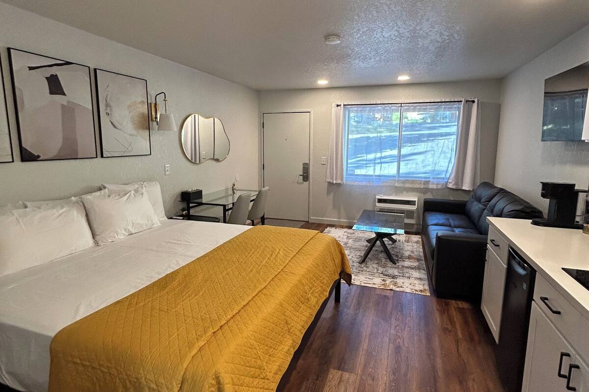 A compact hotel room with a bed, a sofa, a small kitchenette, and a window with curtains.