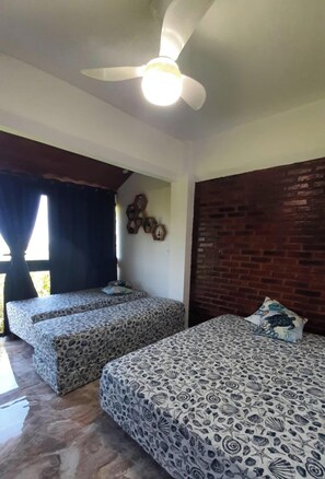 Ein Zimmer mit zwei Betten, einem Deckenventilator, einer Wand aus Backsteinen und einem Fenster mit Vorhängen.