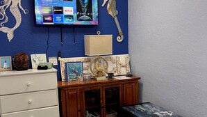 Una habitación con un televisor montado en la pared que muestra varios servicios de streaming, un mueble de madera con objetos decorativos, una alfombra mullida y un cuadro con un faro.