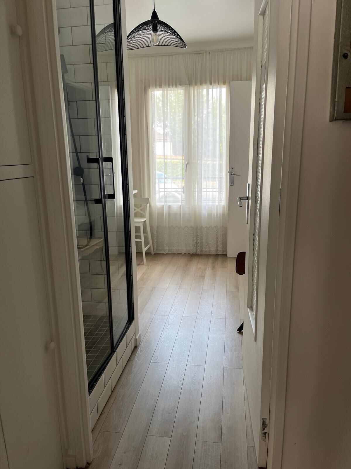 A hallway with a glass door leading to a balcony, a white chair, and a large window with sheer curtains.