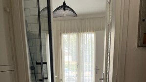 Un couloir avec une porte vitrée donnant sur un balcon, une chaise blanche et une grande fenêtre ornée de rideaux légers.