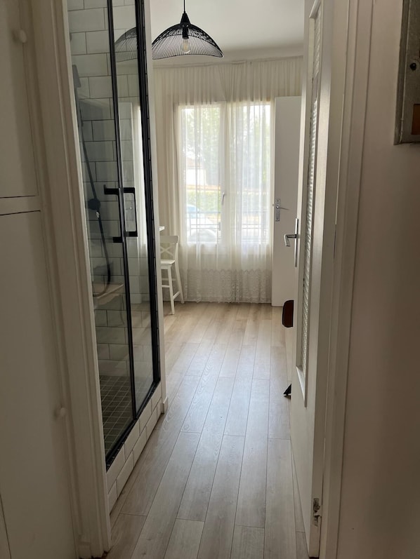 A hallway with a glass door leading to a balcony, a white chair, and a large window with sheer curtains.