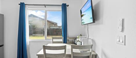 Ein Essbereich mit Tisch, Stühlen, einem an der Wand befestigten Fernseher und einem Fenster mit Vorhängen.