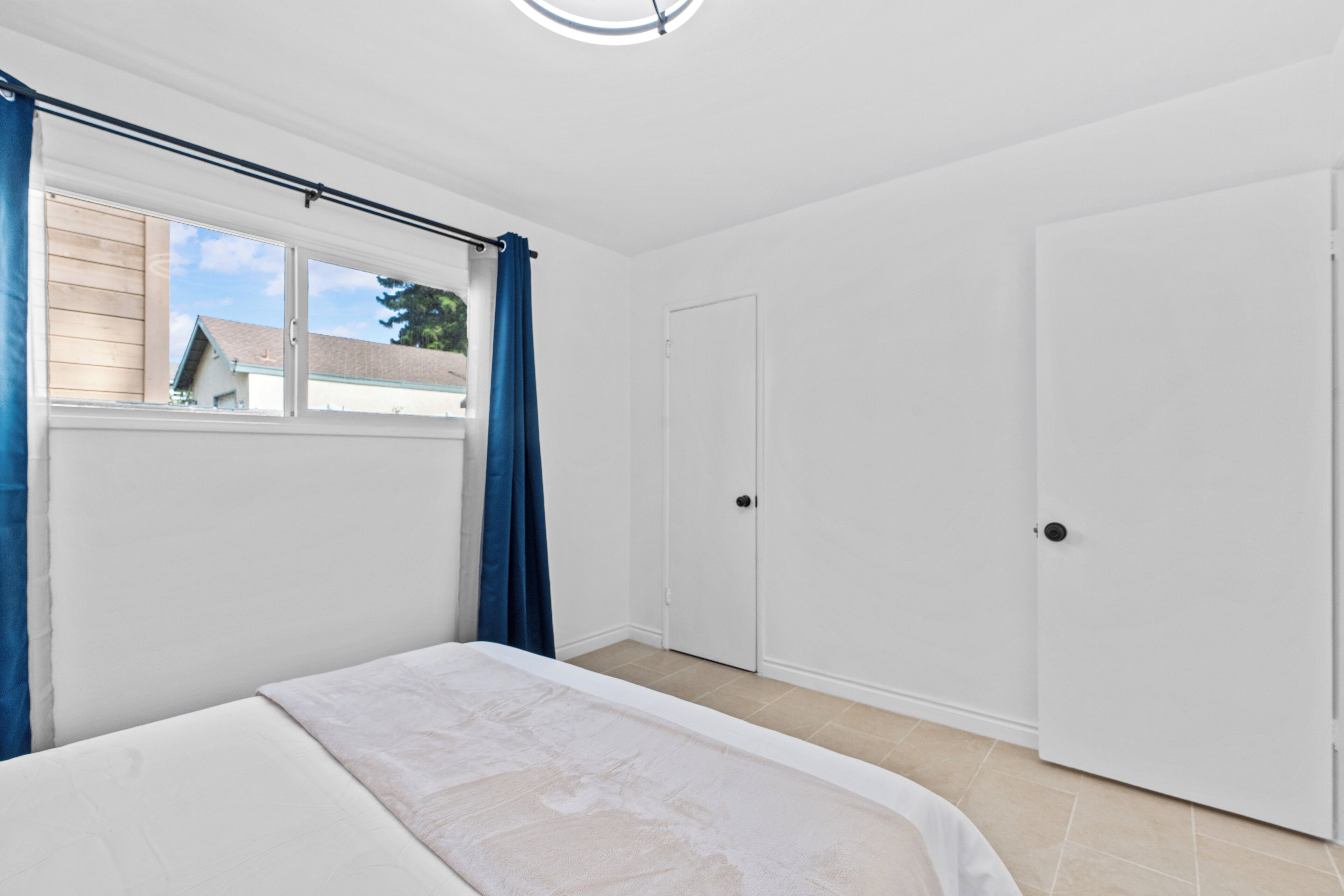 Ein Schlafzimmer mit einem Bett, einem Fenster mit Vorhängen, einer Tür und Blick auf ein Haus.