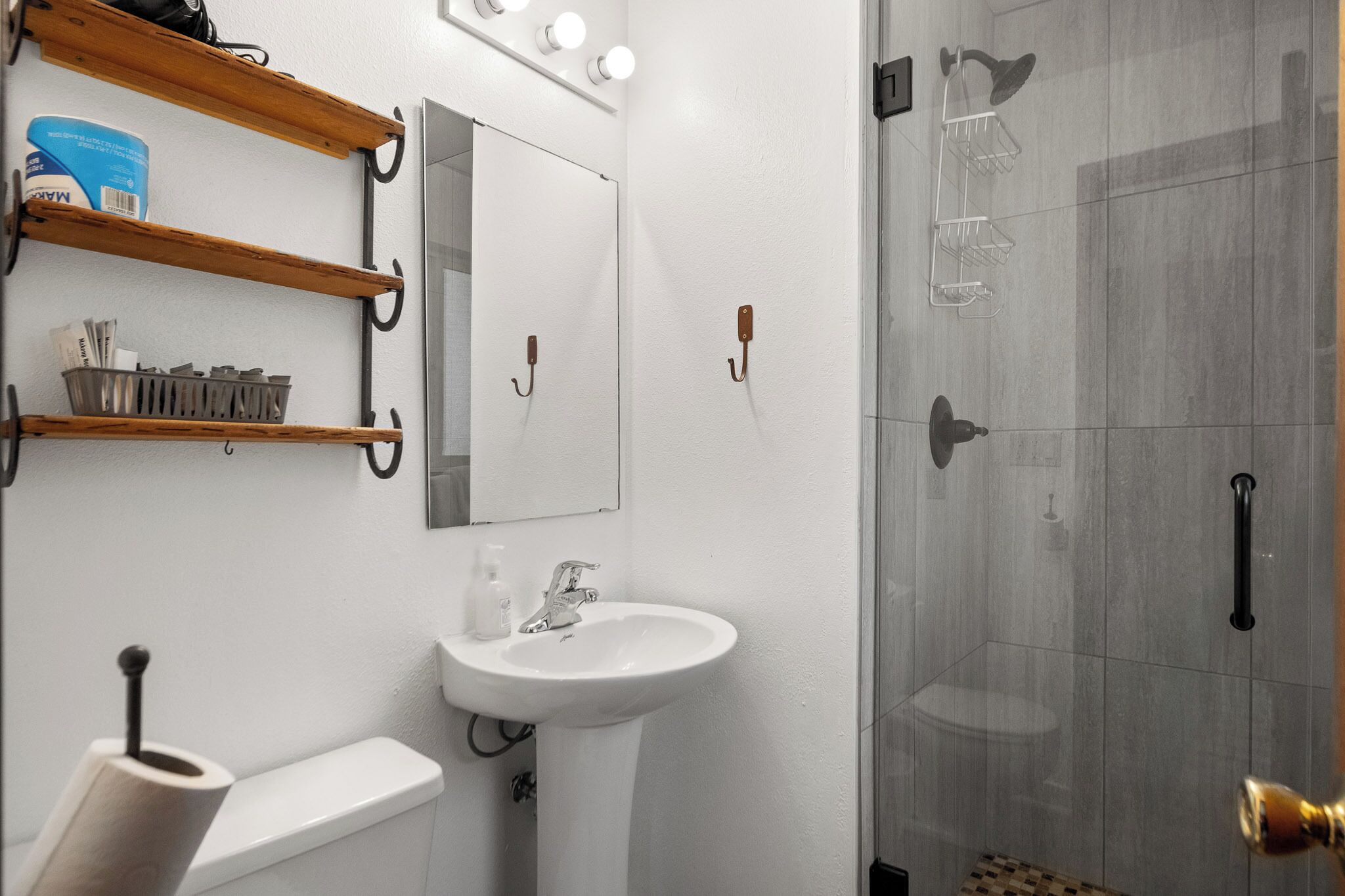 A modern bathroom with a white sink, toilet, and shower. There are wooden shelves above the sink and a mirror on the wall.