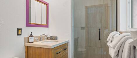 A bathroom with a wooden vanity, a pink-framed mirror, a glass shower enclosure, and a towel rack.