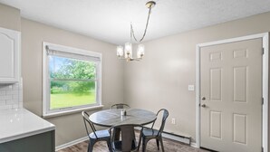 A dining area with a round table, four chairs, a chandelier, a window with a view of greenery, and a door.