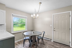 A dining area with a round table, four chairs, a chandelier, a window with a view of greenery, and a door. - Quaint Rennovated 2-Bedroom close to the solar farms and Honda (Raymond)