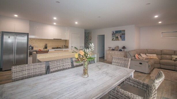 A modern kitchen with white cabinets, a stainless steel refrigerator, and a dining area with a wooden table and wicker chairs. - Azure on Bondi Place (Kingscliff)