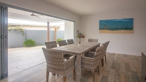 A dining area with a wooden table and wicker chairs, a painting on the wall, and a view of the outdoors. - Azure on Bondi Place (Kingscliff)