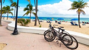 A beach scene with a bicycle parked near a palm tree, a curved walkway, and beach umbrellas in the background. - Fort Lauderdale Luxe - Beach + Rooftop Pools (Fort Lauderdale)