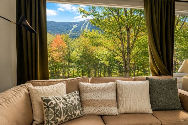 A modern living room with a sofa and patterned cushions, a lamp, and a view of a forest and mountains through a window.