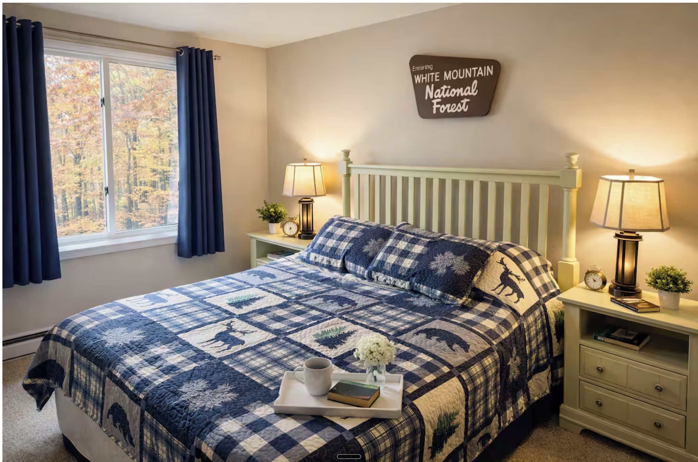A bedroom with a bed, two bedside tables, a nightstand, a window with curtains, a wall sign, and a bedside lamp.