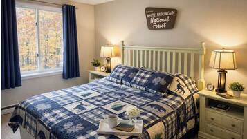 A bedroom with a bed, two bedside tables, a nightstand, a window with curtains, a wall sign, and a bedside lamp.