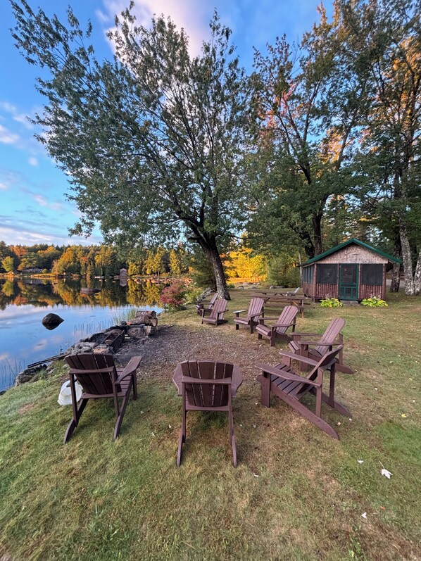 A lakeside setting with wooden Adirondack chairs arranged in a circle, a small cabin, and a natural landscape with trees and a body of water.