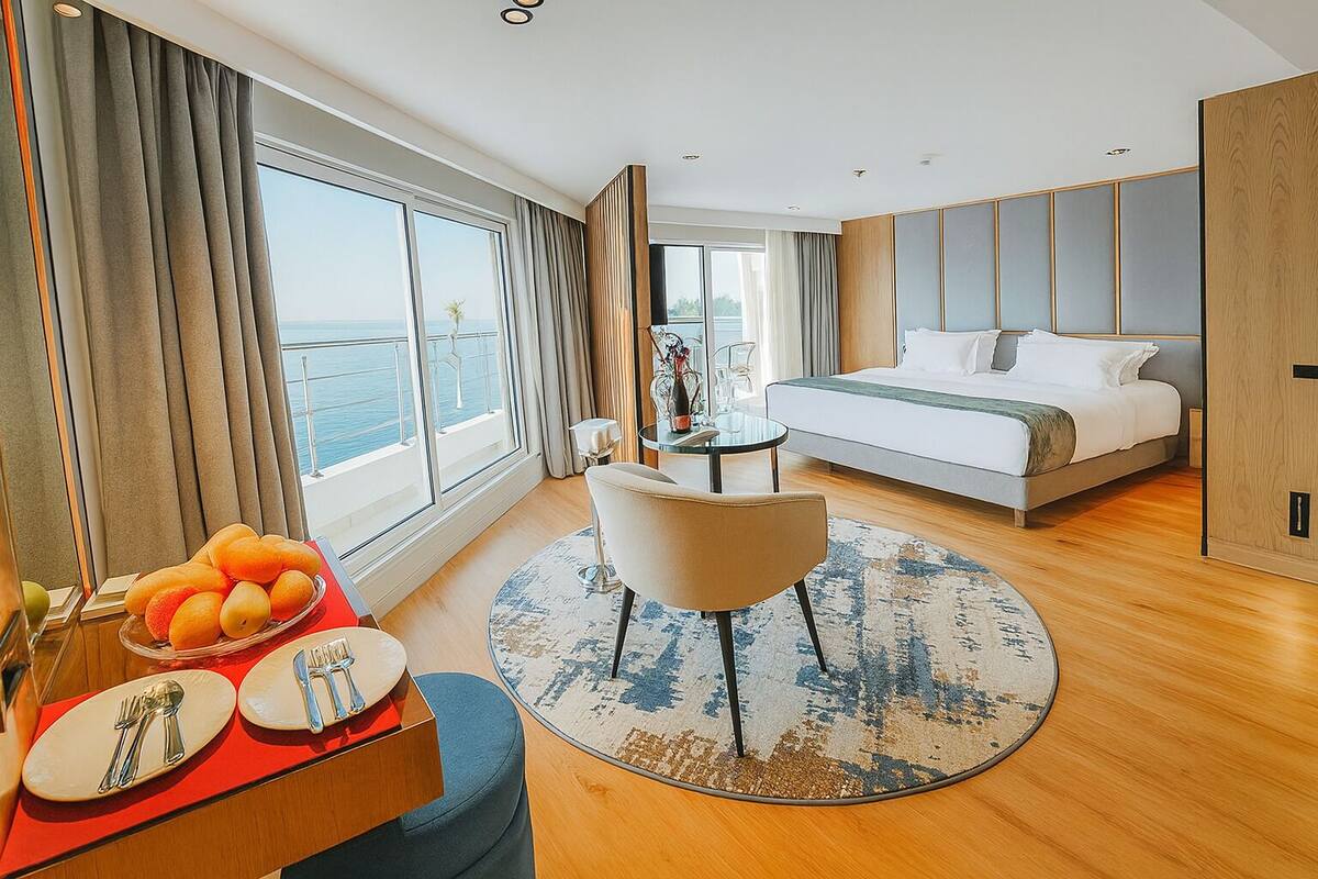 A modern hotel room with a large bed, a round dining table with a chair, a fruit bowl, and a view of the sea.